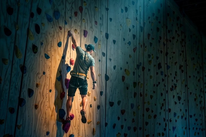 Person climbing indoor rock wall with colorful holds, spotlighting from above.