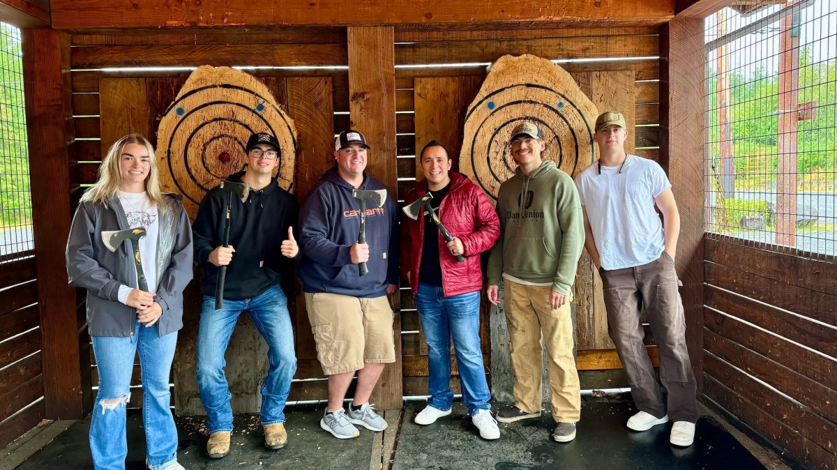 Group of six people posing in front of wooden axe targets, holding axes.